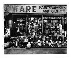 View of a hardware store display, with an assortment of tools and other objects in the window and displayed on the sidewalk in front of the store.