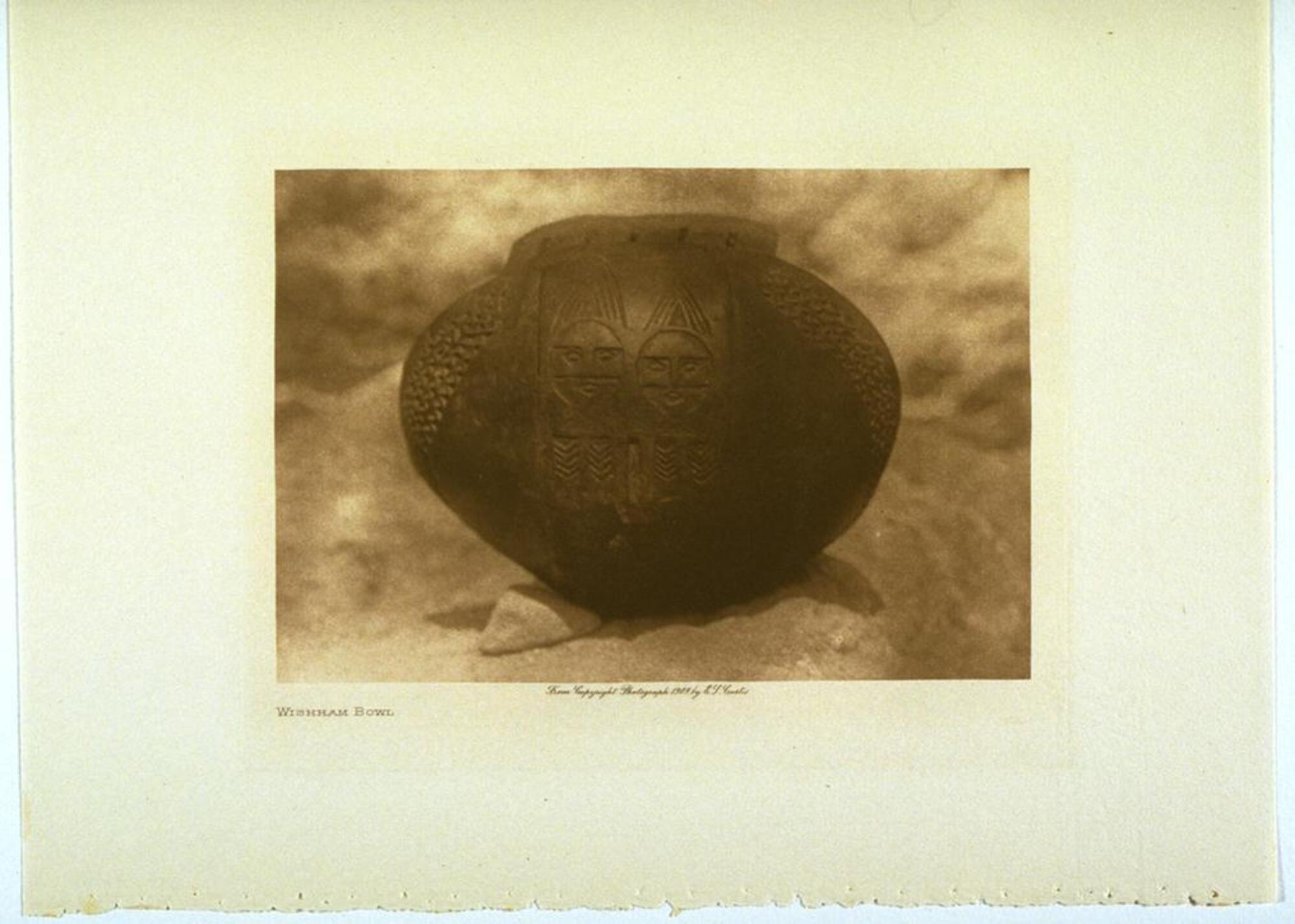 A photograph of a hand-made ceramic bowl placed on a desert rock. Two abstract human figures are etched into the surface of the bowl, with decorative textures carved into the surface on either side.