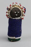 Fabric cylindrical body with round fabric head. Beaded blue body with white and green detail at bottom. Darker fabric face with white beaded detail. "Hair" in large white beads and small red beads. Blue beaded handle attached to head, white tag attached.