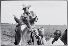An officer on top of a horse wearing a white cowboy hat. There is an African-American man standing next to him handing him water.
