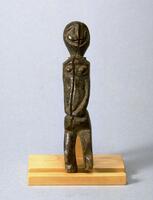 Standing human figure with arms in front of the body and slightly bent knees. The head is round with minimal detail. There is a large crack through the center front of the figure. 