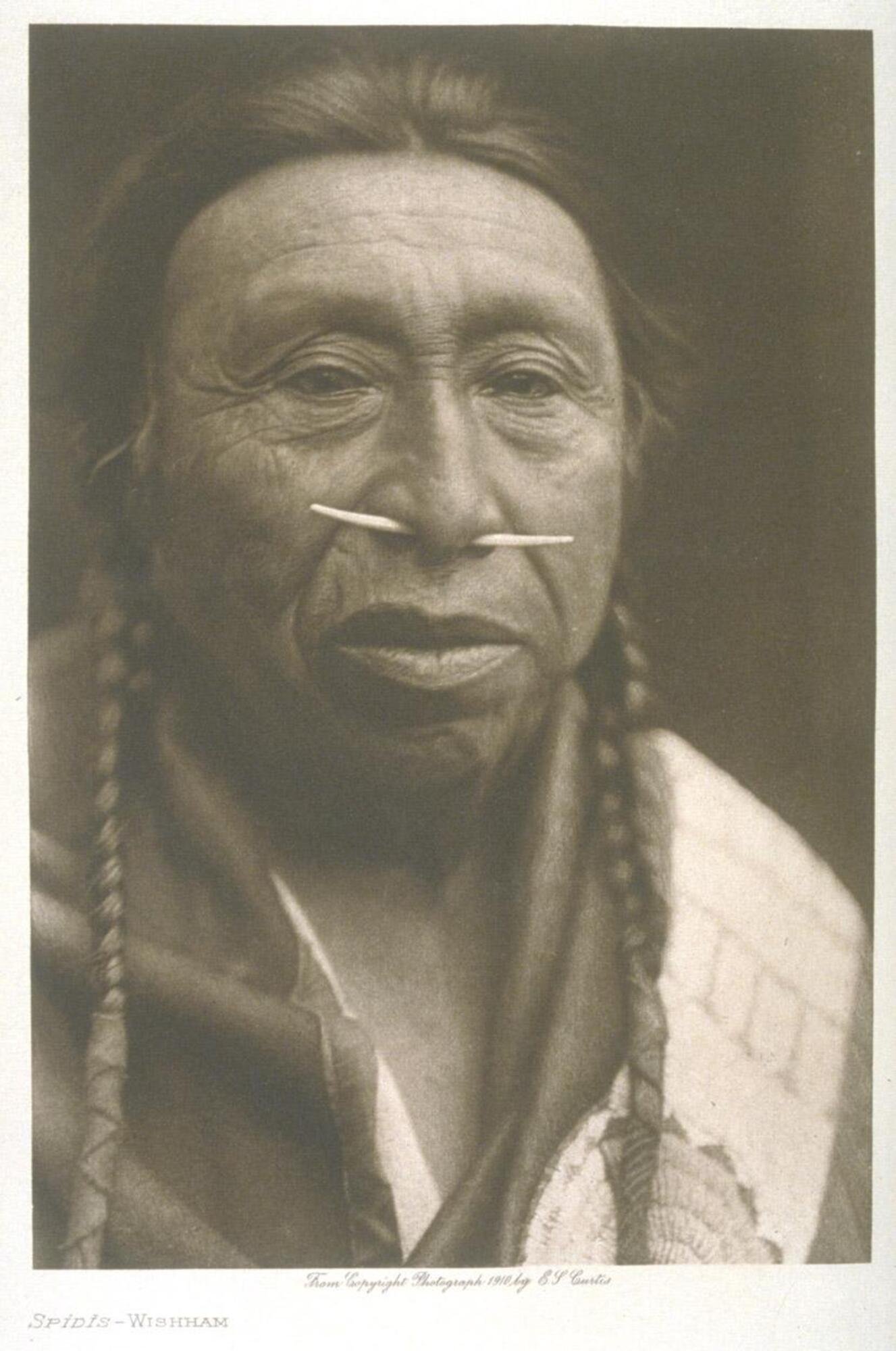A portrait of an aging man. He wears a tusk septum piercing and two long braids, with a garment draped over his soulders. A circular embroidered design is visible near the bottom of the frame.