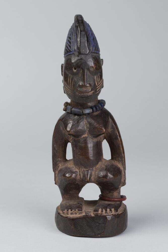 Standing human figure on a round base with hands at the sides. There is a metal loop around one ankle and a string of black beads around the neck. On each cheek are four horizontal marks topped by three vertical marks. The hair is in a conical shape with a raised central groove surrounded by diagonal lines on each side and coated with blue pigment. 