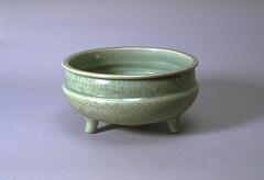 This is a round bottomed bowl on three legs with high straight sides and and everted rim. It is covered in a green celadon craqueleur glaze.