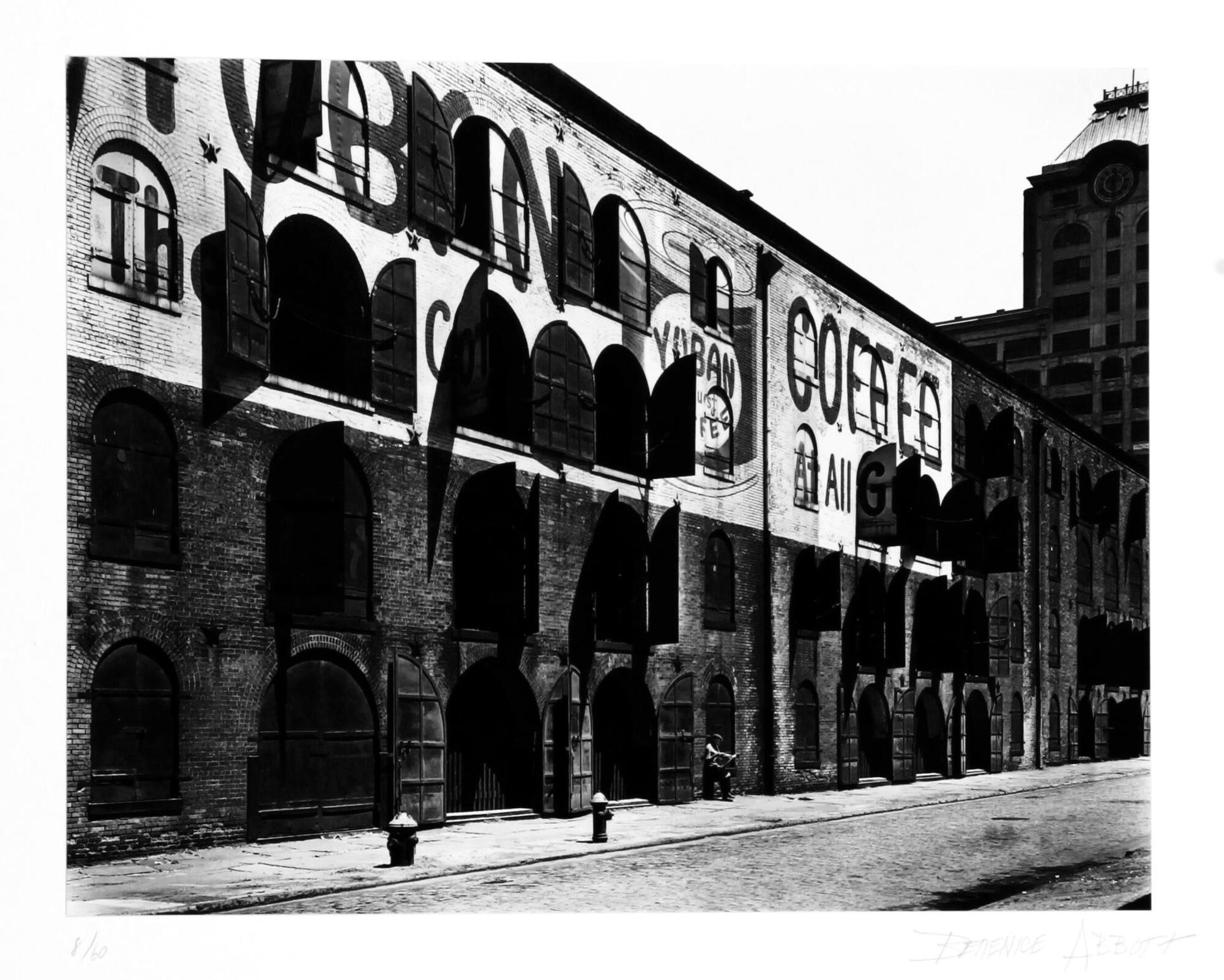A black and white photograph of a warehouse in Brooklyn. A sign painted on the building advertises coffee, while a man sits outside reading a newspaper.