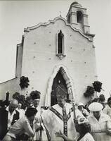 A clergyman dressed in robes and a tall, pointed hat smiles and reaches his right hand to a woman who bends to kiss it. Several other men and women surround them, all standing in front of the simple facade of a tall building.