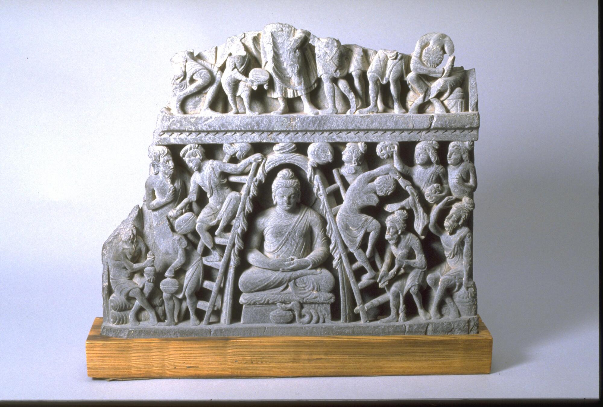 A fragment of a stone slab, originally a facing on the drum of a small stupa, carved with a narrative scene.  In this relief, he is converting devotees of the fire god Agni, the three ascetic Kasyapa brothers and their disciples. In this narrative, the Buddha asked to spend the night in the fire temple of Uruvilva Kasyapa. The temple’s fire god worshipers thought the fearsome fire serpent that dwelled within the temple would vanquish the Buddha.  Instead, the fiery radiance of the meditating Buddha overwhelmed the serpent, who crawled into the Buddha’s alms bowl; the defeated snake appears in this sculpture below the Buddha. Meanwhile, the dazzling radiance the Buddha emits has been mistaken for flames and a fire brigade using ladders and pots of water has been formed to put out the fire, as can be seen here. Seated in the posture and gesture of meditation, the Buddha’s calm presence is in contrast to the action unfolding around him. The three Kasyapa brothers, with their beards and matted hair, are at the bo