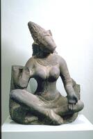 Varahi has a crowned boar’s head on a woman’s body.  She sits with her ankles crosses and originally had four arms, the back two have broken away as has the front right hand which had probably been held up in a reassuring gesture.  Her left from hand is held down at her left knee with the palm held facing out in a gesture of giving.  Her body is softly modeled with a narrow waist and full breasts.  She wears jewelry that is in sharp but low relief and includes a series of necklaces forming a collar of decoration and a longer one that falls between her breasts which suggests the sacred thread extending down to her waist at her right.  She is naked from the waist up and the lower garment is merely suggested by the heavier folds at the waist.  Her head is tilted and her chin/snout had jutted out to the left, but the lower snout is broken away.  She wears a conical crown that accents the long diagonal of her face.  Originally there was an arch behind the image which would have supported her back arms, so the imag