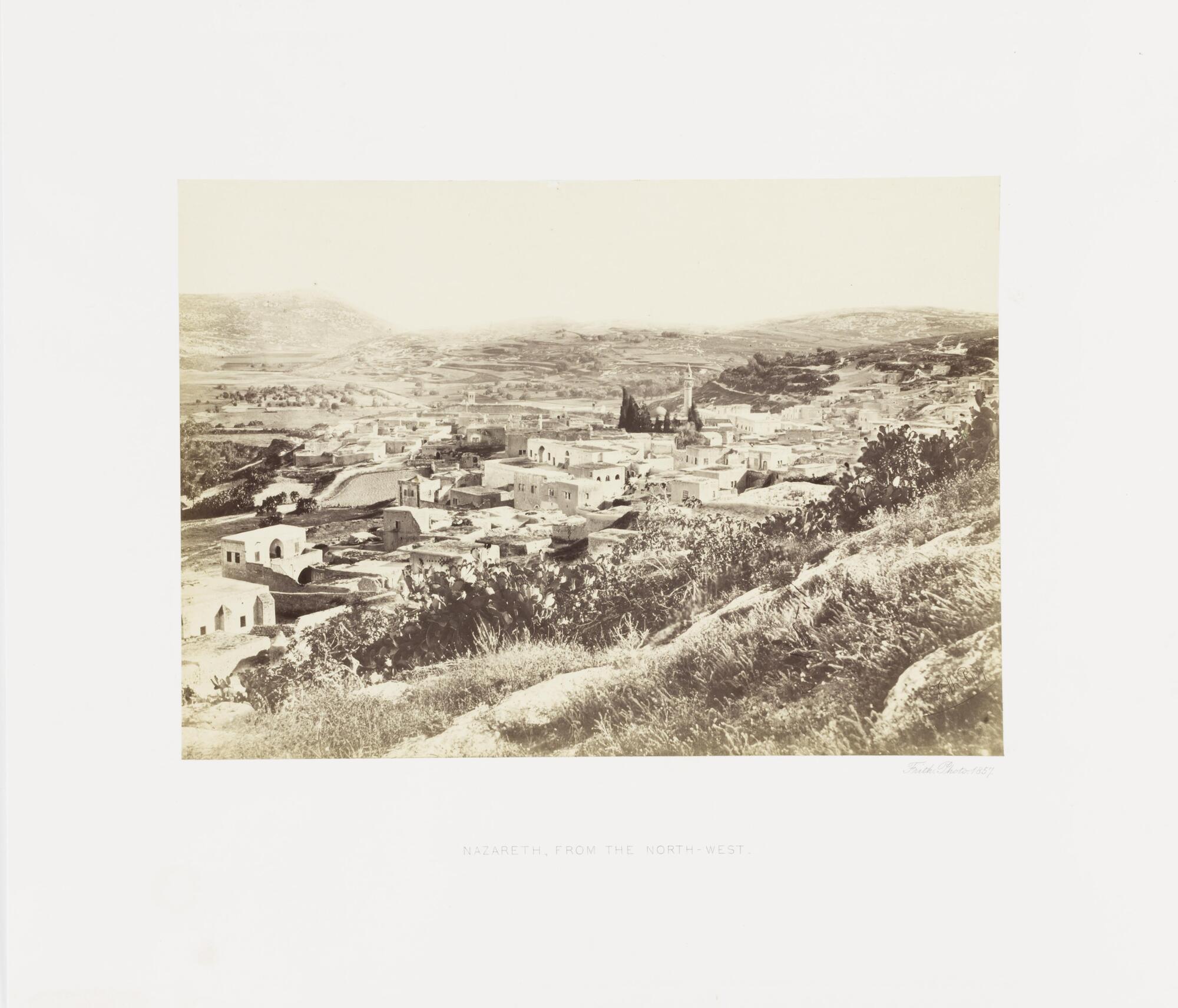 View of a village set in a valley surrounded by several low-rising hills, all of which is seen from an elevated perspective from a hill in the foreground of the image. 