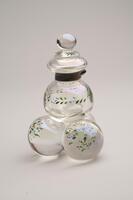 This is a clear glass inkwell with a metal collar and sphere finial lid. The body has a hollow glass sphere resting on a base of three solid glass spheres. Each of the spheres is decorated with painted leaves and flowers.<br />