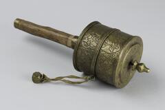 A &#39;mani&#39; or prayer wheel, a common ritual object used in Tibetan Buddhism: a device made a handle (here a simple wooden stick) supporting a hollow cylindrical drum on a spindle. A small metal weight, attached to one one side of the drum with a cord, allows the wheel to spin with a slight rotation of the wrist. The drum in his case is of hammered metal, with incised patterns of Buddhist auspicious symbols.