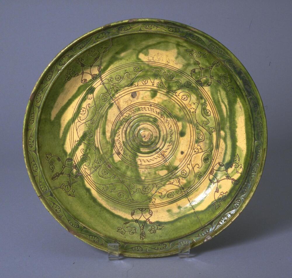 The dish belongs to a large group of sgraffiato wares, examples of which have been found from Afghanistan to northwest Iran. They are characterized by an incised design cut into a slip and enhanced with glazes of different colors, frequently yellow and green. In this particular case, and others like it, the concentric scratched lines are clearly determined by compass while the filler patterns are somewhat less controlled. The pigment is not applied to coincide with the engraved line but rather forms an independant web of color over it.<br />
 
