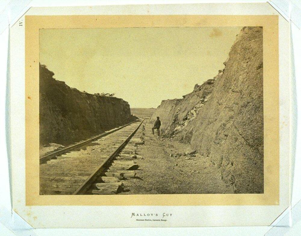 This photograph depicts railroad tracks cutting through a man-made passage.  To the right of the tracks stands a lone man looking into the distance.