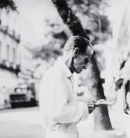 This photograph depicts the profile of an elderly man dressed in a white shirt in an urban environment. The man's hands are extended before him and appears to be counting coins.
