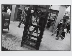 This photograph depicts a view of a city sidewalk with pedestrians walking in front of a bar. In the middle of the frame is a phone booth with a woman inside, talking on the phone.