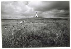 Photograph of field with wild flowers. Church is visible in background. Dark, thick clouds loom overhead.<br /><br />
Eva Caston 2017