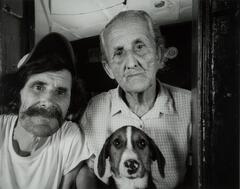 A portrait of two men and a dog. The man on the left is wearing a hat and white shirt; he has a large moustache. The older man to the right is wearing a plaid button down shirt.