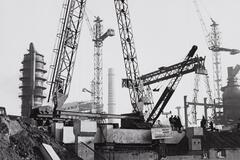 Photograph of a construction site with numerous cranes, silos, and smokestacks. Three men confer in the lower right.