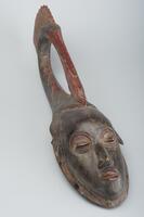 Wood mask of an elongated face topped with a bird with red plumes.
