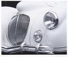 Front end of a classic car showing one headlamp. The car is very light in color and the features are chrome.