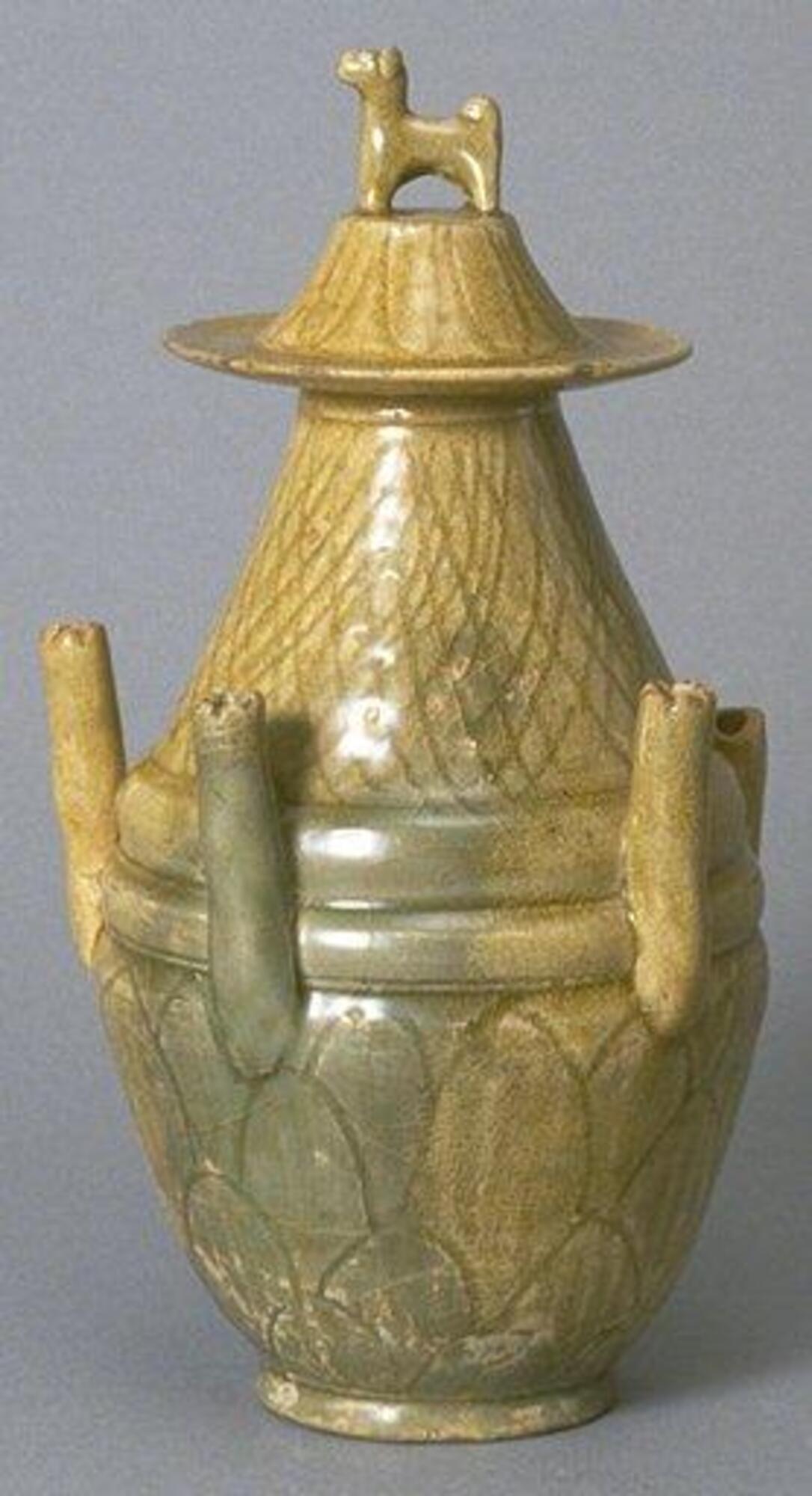 A buff stoneware jar rising up from a tall foot ring in an elongated globular body with lobes tapering towards the mouth.  There is incised decoration, and five tubes evenly spaced and protruding upwards from the belly. The mouth is covered with a high truncated conical lid, incised, and topped with a dog finial.  It is covered in an olive green celadon glaze.  On the lid is accession number 1987/2.46B.