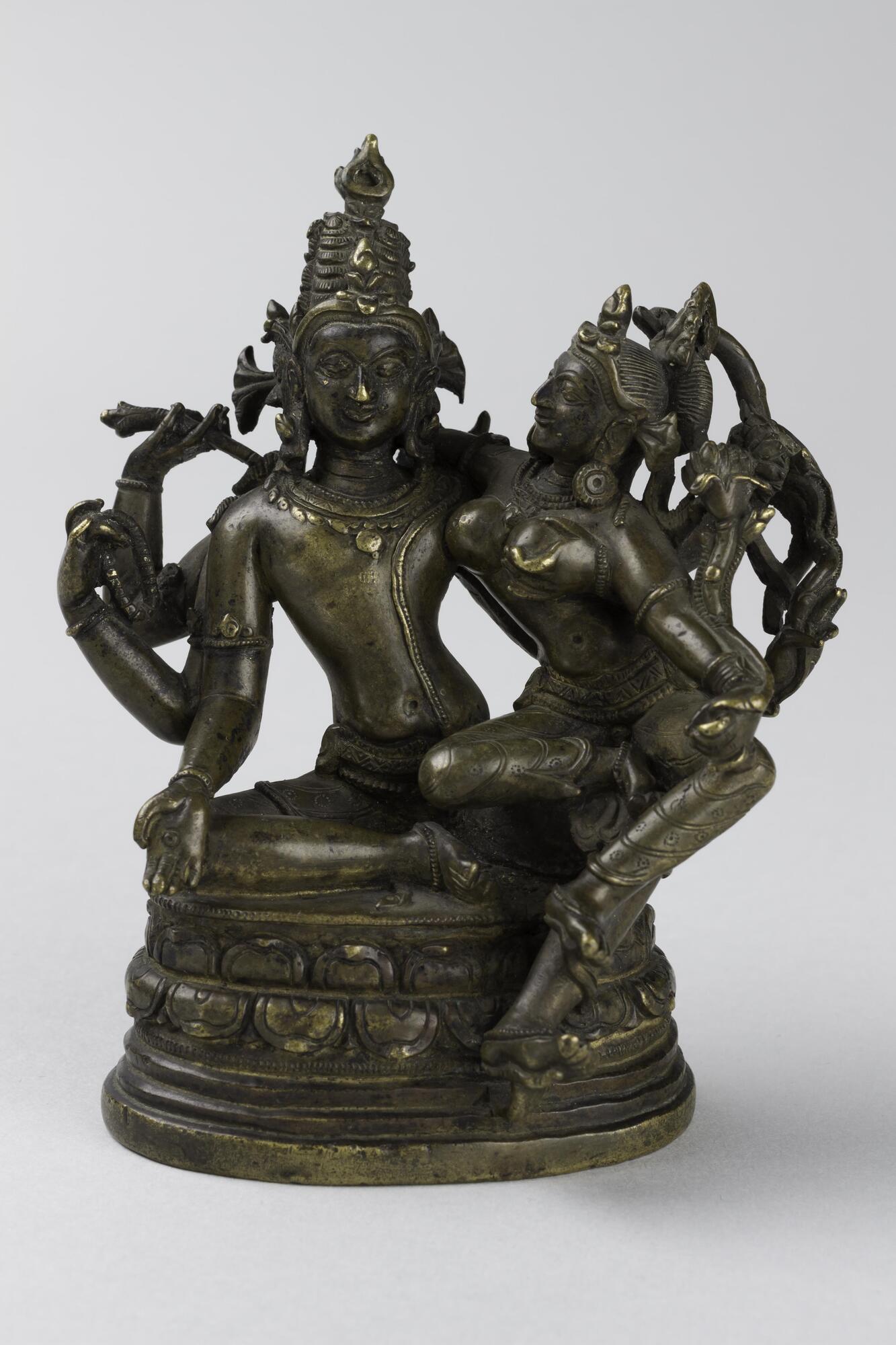Shiva sits with his consort on a double lotus pedestal.  He has six arms, his right three are in varada mudra [a giving gesture], holds a rosary and an arrow.  His left arms cup his consorts left breast and hold a lotus flower and a bow.  He sits in royal ease, with one leg pendant.  He wears bracelets, armlets, necklaces, earrings, and a sacred thread that stretches form his left shoulder down past his waist.  On his head he wears an elaborate jatamukuta, a crown interlaced with his matted locks.  Parvati sits upon his knee with one leg tucked under her and the other pendant.  She is also adorned with jewelry, but wears a more modest diadem at the front of her head.<br />