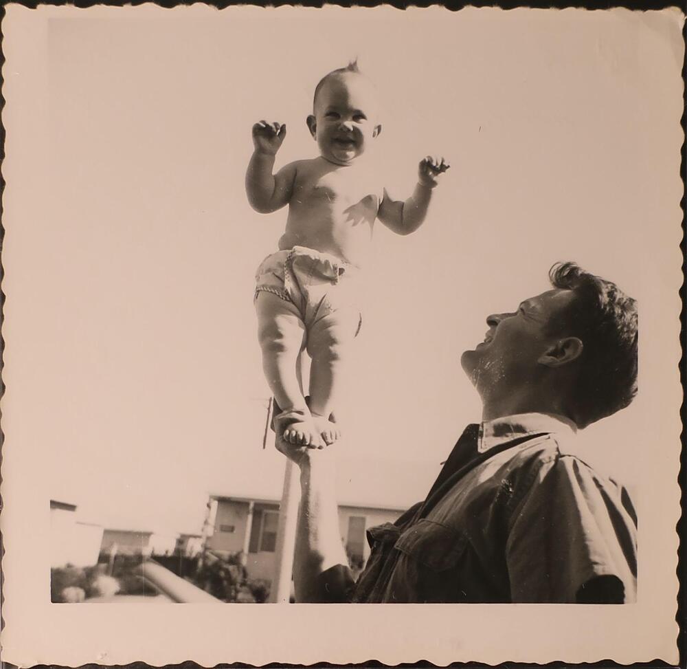 A man balancing a smiling baby in his right hand. The man holds the baby&#39;s feet and the baby is standing upright in the center of the image.