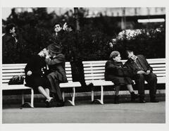 Image of two older couples sitting on separate benches. The left man and woman hold cigarettes and kiss; the right woman looks off to the left while her partner sleeps. Four dark-haired young men stand behind them.&nbsp;