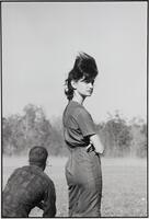 A photograph of a man crouching and a woman standing in a grassy field. The man bends over with his back toward the camera. The woman turns her body toward the right side of the frame, looking into the distance as her hair blows in the wind.