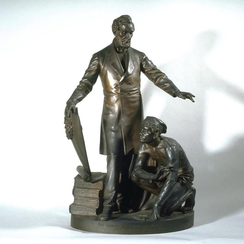 Bronze sculpture of a standing male figure his right hand holding a shield which rests upon a stack of book while his left arm is outstretched hovering over the crouching figure of an African American male figure.