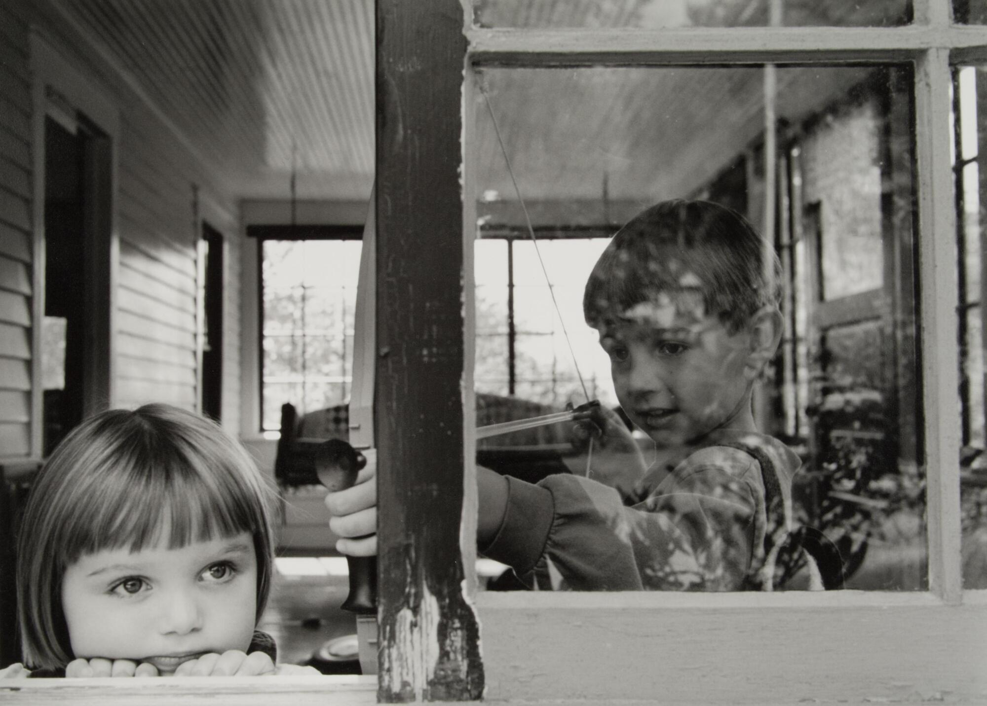 This photograph is of two children in an enclosed porch. A young girl rests her head on a ledge, while a young boy stands behind her to the right, holding a toy bow and arrow close to her head.