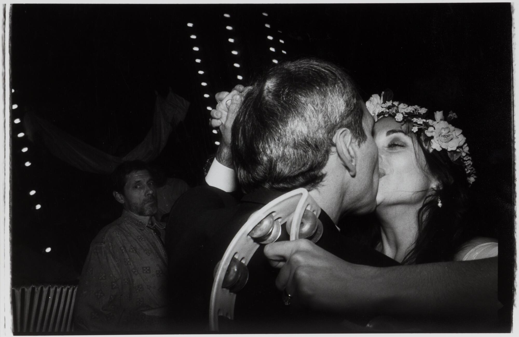 A photograph of a man and woman kissing; the man's head turned from the camera. The woman holds a tambourine in her hand and wears a flower crown. In the background, a man looks toward the camera.