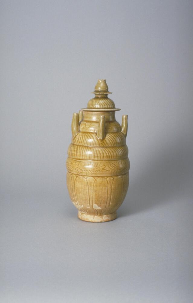 This buff stoneware jar rises up from a tall foot ring in an elongated globular body with lobes tapering towards the mouth. It has incised decoration and five tubes evenly spaced, protruding upwards from the shoulder. The mouth is covered with a high domed lid, incised, and topped with a lotus bud finial. It is covered in an olive green celadon glaze.