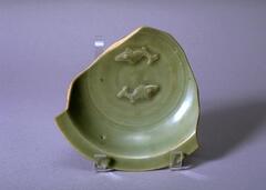 A small stoneware bowl on a foot ring with everted flat rim.  The interior is molded with two fish and covered in a green celadon glaze.  There is significant loss to rim and sides.