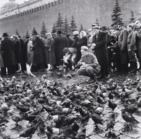 A young soldier crouches down amid a crowd of people to feed a large flock of pigeons. 