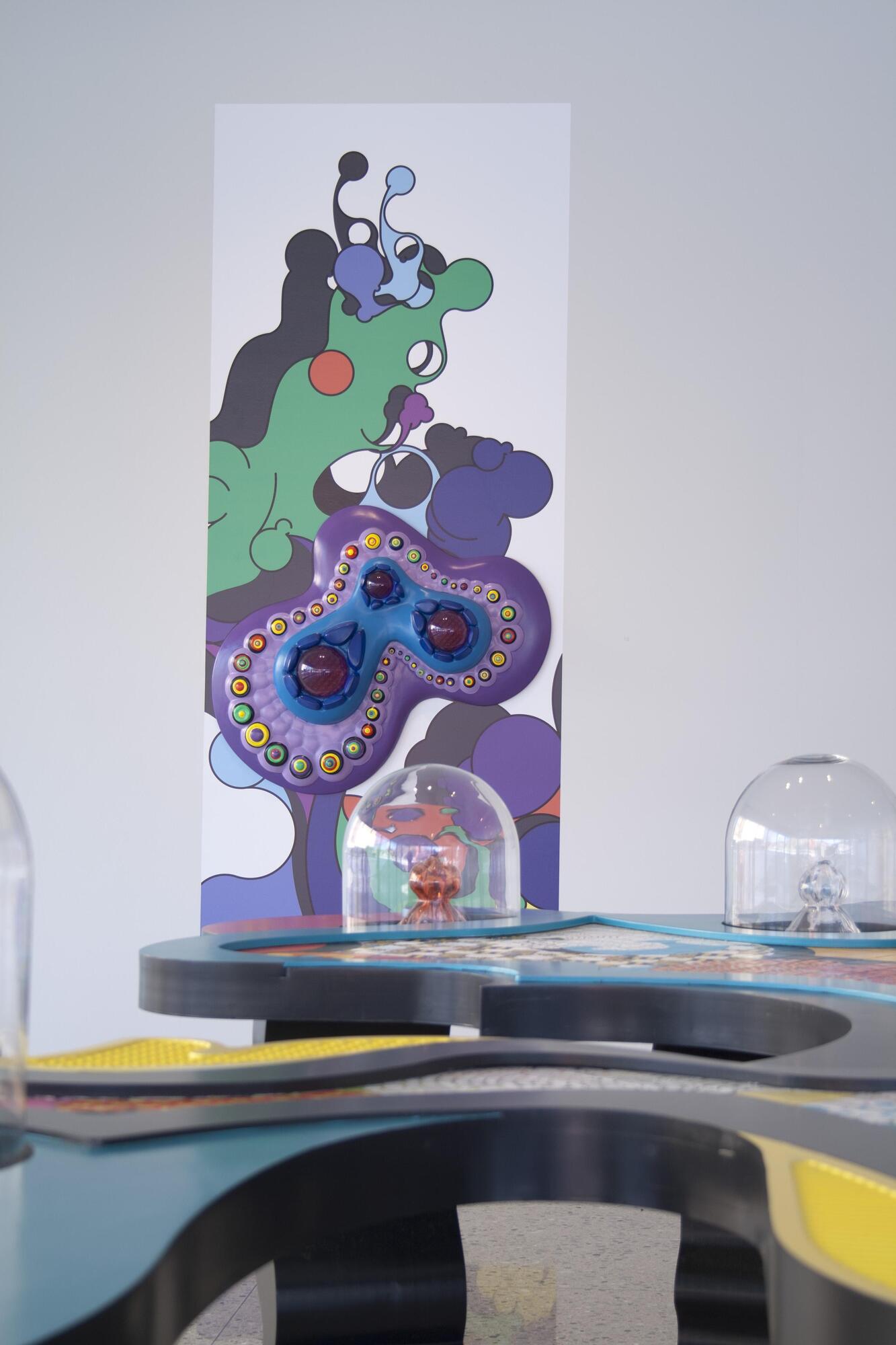 A purple omeba-shaped form that has mutli-colored circles and blue designs on top. It hangs on a wall on top of a vinyl rectangle that has similar designs.