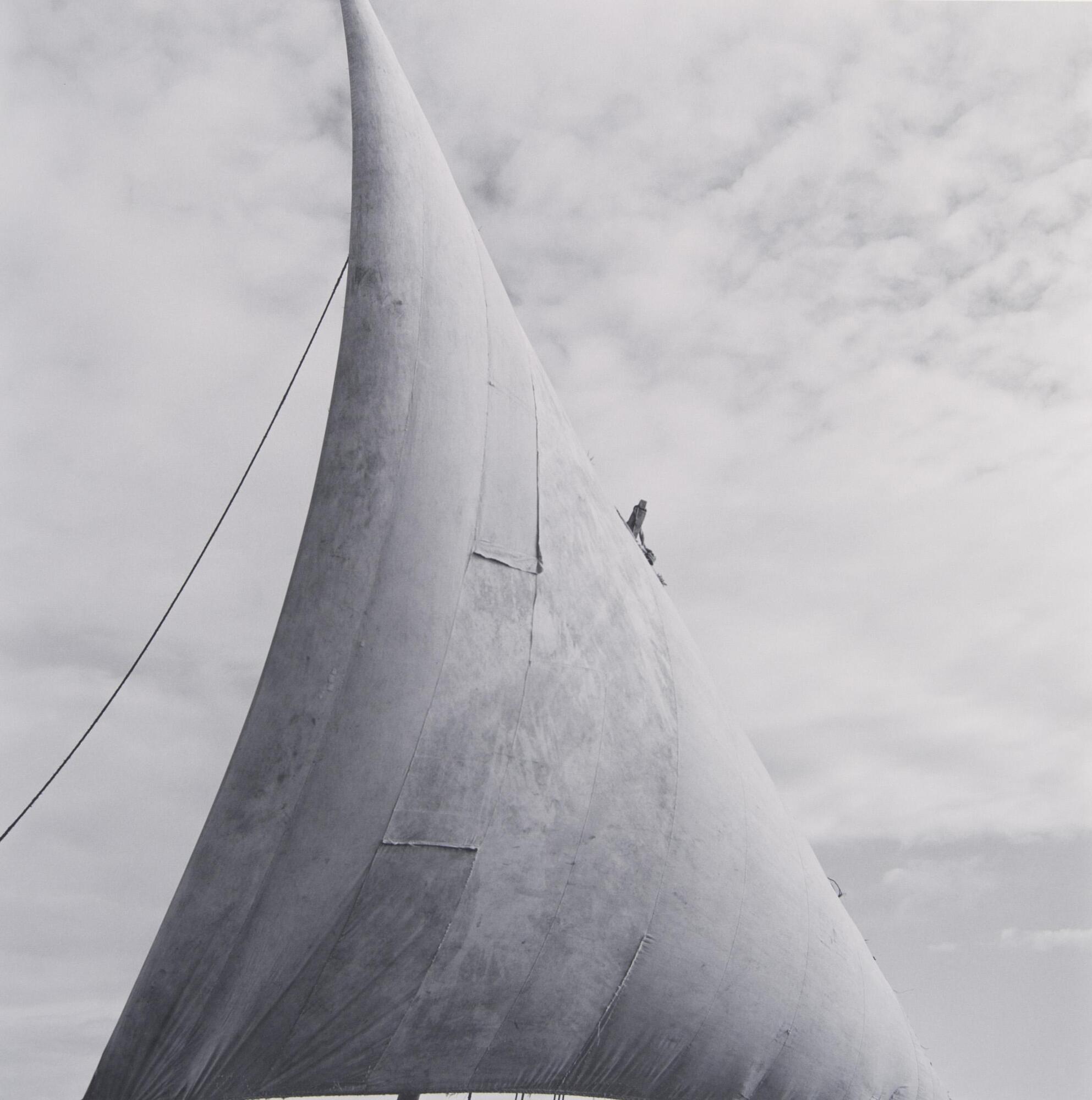 A large ship sail against a cloud-filled sky in Kenya. The angle from which the photograph was taken creates a triangular shape to the sail. From the top corner to the left edge of the photo is a rope, and on the right side of the sail is a small black figure protruding from behind the cloth, possibly the top of the mast to which the sail is connected.