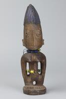Standing female figure on a round base with hands at the sides. The figure has prominent breasts and strings of multi-colored beads around the waist and neck. The pupils of the eyes are metal and the hair is in a rounded comb shape with vertical grooves and blue pigment. 