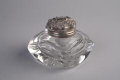 A crystal inwell with a cube body and rounded edge corners and a silver lid on top.