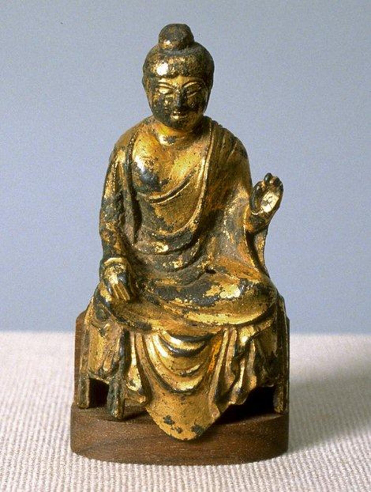 Small gilt bronze seated Buddha in style of China’s cosmopolitan Tang Dynasty. It has a full figure, artfully draped robes, and a plump, rounded face with arched eyebrows.   Hand is raised in abhaya mudra.