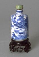 A porcelain snuff bottle with blue underglaze. Painted on the bottle is a landscape scene of houses with people inside, trees, a body of water, and a man standing on the shore. On the top is a green jadeite snuff bottle.