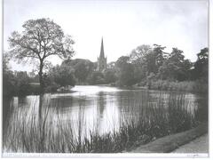 This photograph shows a pond which is lined with grass and trees. A church with a tall steeple is in the background, at the waters edge.