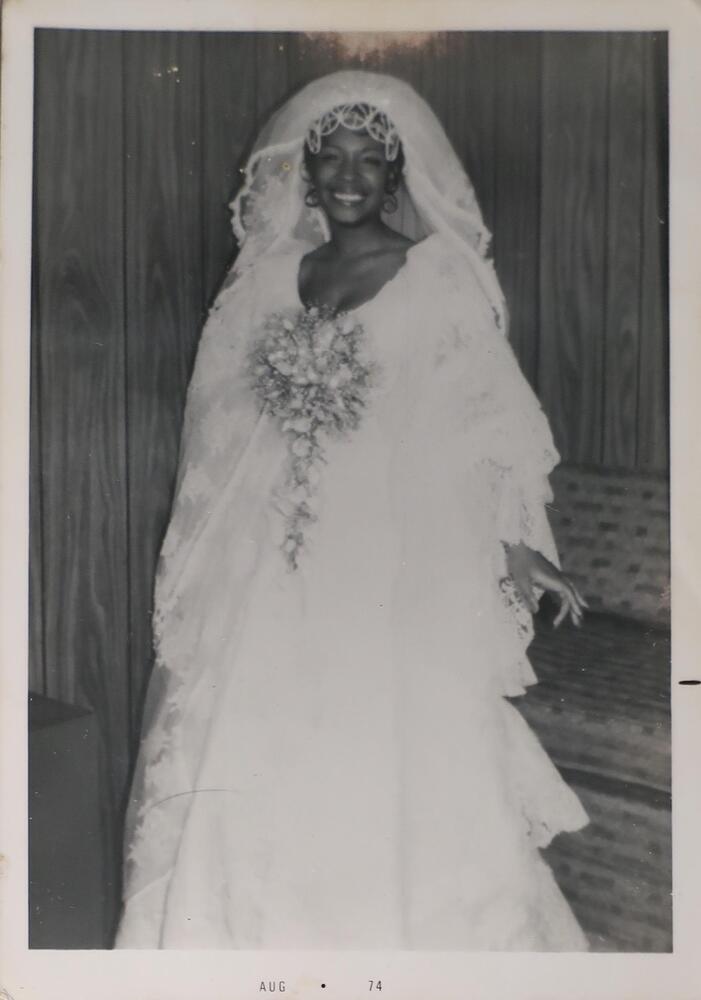 <span style="font-size:12pt"><span calibri="" style="font-family:">A portrait of a smiling woman in a wedding dress and veil with a bouquet of flowers held at her chest. Her left hand is extended slightly behind her. She is standing against a backdrop of mostly wood paneling.</span></span><br />
&nbsp;