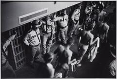A group of prisoners, naked, standing in a line next to a line of officers. They are in the hallway between the cells.