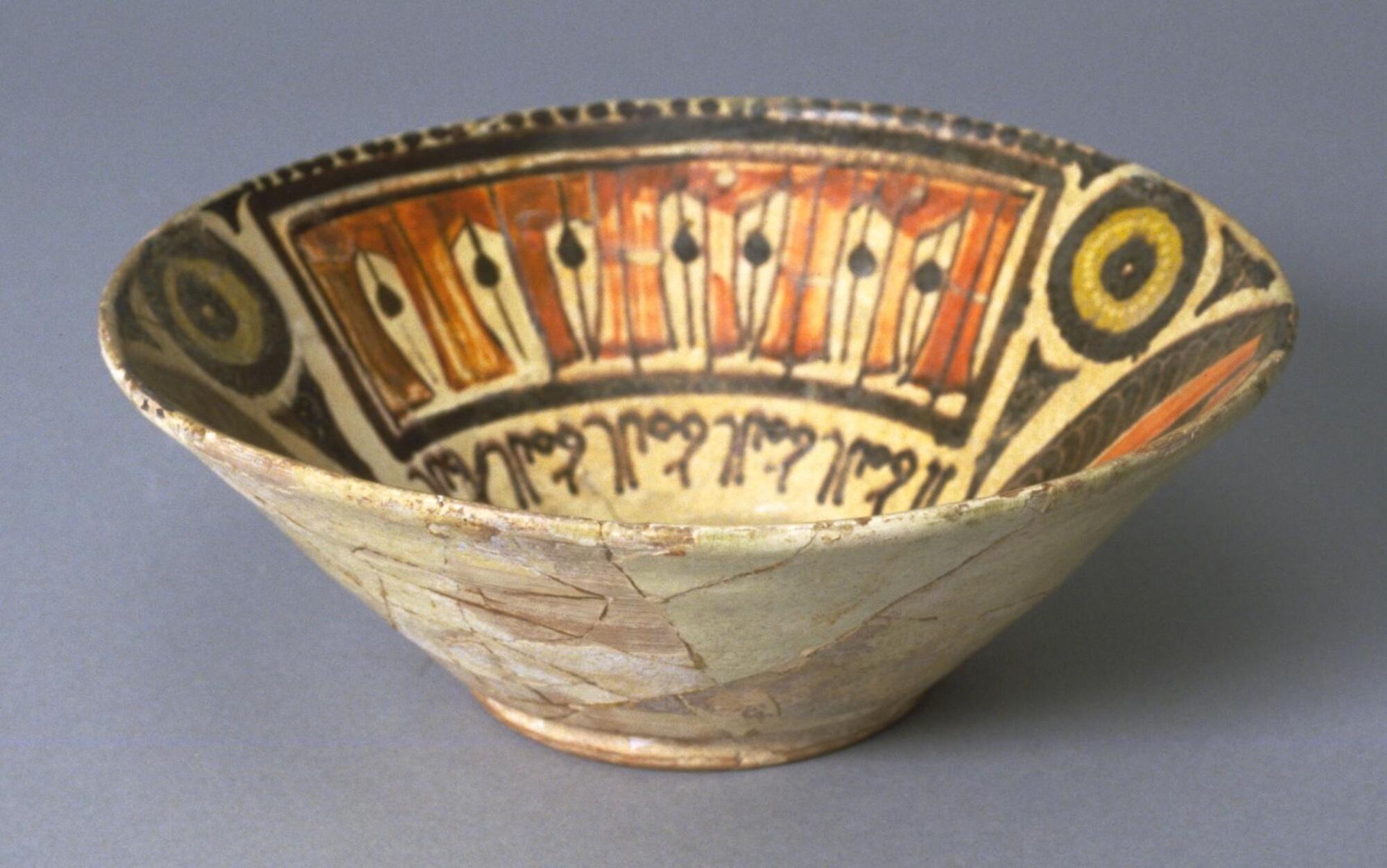 The vibrant coloring of this deep bowl consists dark brown, muddy brown, yellow-green and ivory. The design is organized around an inscription which appears above the base far down on the side. Above the inscription running horizontally is a separate enclosed design which looks like arcaded doorways with a string with a ball on it cutting each arcade in half for a total of seven. Opposite this is a series of four and one-half arcades in a vertical direction. Between these two areas there is a design repeated on opposite sides of the bowl with floral circular motifs on each side of a large, abstracted pear-shape form. The circles are brown and yellow-green and the pear form is orange and brown. Beneath each circle is a 'base' narrowing towards the center and directing the eye to the base. The base is covered with a shield-like design of orange and brown. The ivory background is not filled in but certain lines have been incised to add greater interest.  <br />
 