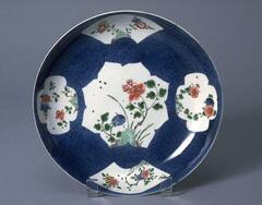 The interior of this thin porcelain, wide, and low saucer-shaped dish on a footring, is covered with sprayed powder blue underglaze with one large foliate-shaped reserve flanked by two smaller fan-shaped reserves and two smaller quatrilobed reserves, and covered in a clear glaze. All the reserves are painted with overglaze enamels to depict peonies, chrysanthemum and prunus sprays, the white exterior is painted with floral sprays, with underglaze blue double ring with central <em>ruyi-</em>shaped <em>lingzhi </em>mushroom mark to the base.<br />
Part of a ten-piece garniture set which includes: jars, 1982/1.206A, 1982/1.206B, 1982/1.206C, 1982/1.207A, 1982/1.207B, and 1982/1.207C; vases, 1982/1.208, 1982/1.215, 1982/1.216, 1982/1.220; plates, 1982/1.212, 1982/1.213, and bowls 1982/1.221,1982/1.22.
