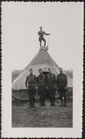 Four men in uniform stand in front of a pyramid-shaped tent. A fifth man is standing at the very top of the tent holding a broom.