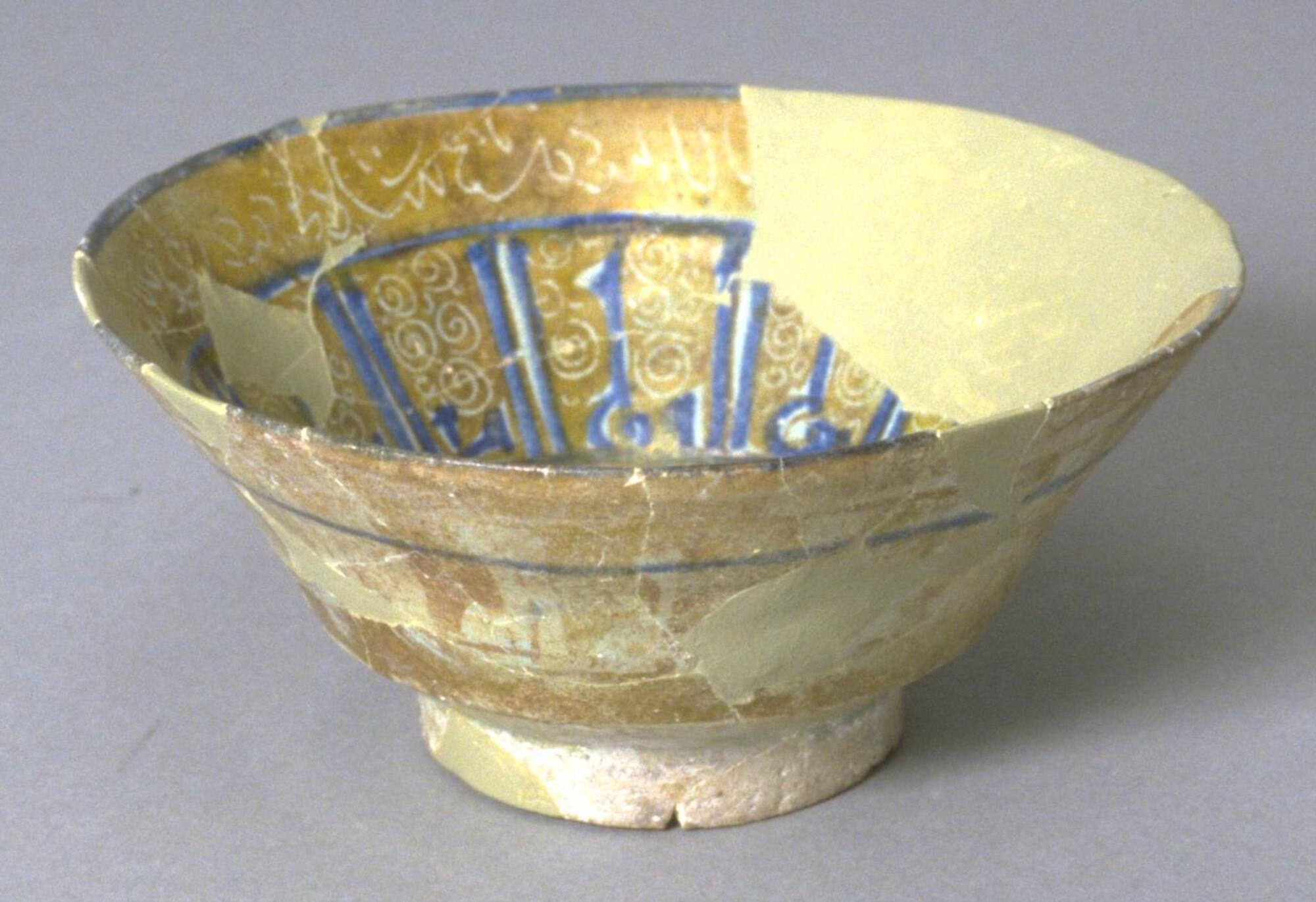 This <em>Kashan</em> style bowl has flaring walls and is made with opaque turquoise glaze, yellow-brown lustre painting and blue overglaze painting. The bowl contains Kufic inscription in cobalt which encircle the interior, with white inscriptions around the rim and center. A bird motif decorates the interior base.  On the outside, we find white Kufic inscriptions under a blue cobalt line encircling the upper part of the bowl.<br />
 
