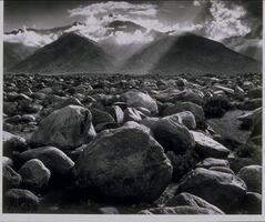 This photograph depicts a flat, boulder-strewn valley leading up to a range of cloud-swept mountains in the distance. 