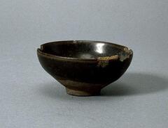 This deep, conical bowl on a straight foot ring with a subtle rim articulation is covered in a thickly applied, dark iron-rich black glaze with subtle hare's fur (兔毫盏 <em>tuhao zhan</em>) markings. There are chips to the rim. 
